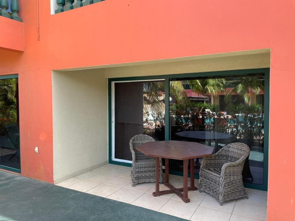 Un área de estar al aire libre con una mesa y dos sillas de mimbre, un puerta de vidrio y vistas a palmeras y un edificio al fondo.