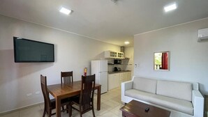 A living room with a sofa, a coffee table, and a dining table with chairs. There is a kitchen area with cabinets and appliances in the background. - Del Rey Apartments (Noord)