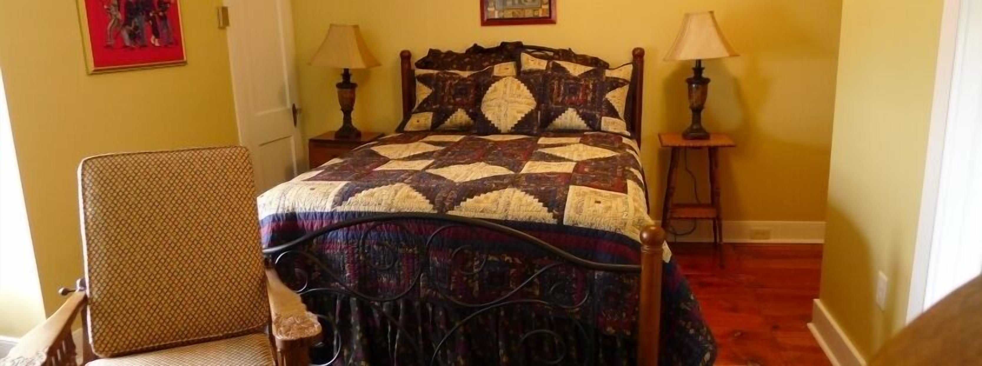 Una cama con colcha estampada, dos lámparas de noche y una silla con asiento de fibras trenzadas.