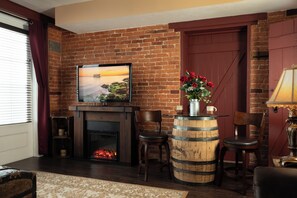 A cozy living room with a brick wall, a fireplace, a television mounted above it, and a wooden barrel table with chairs.