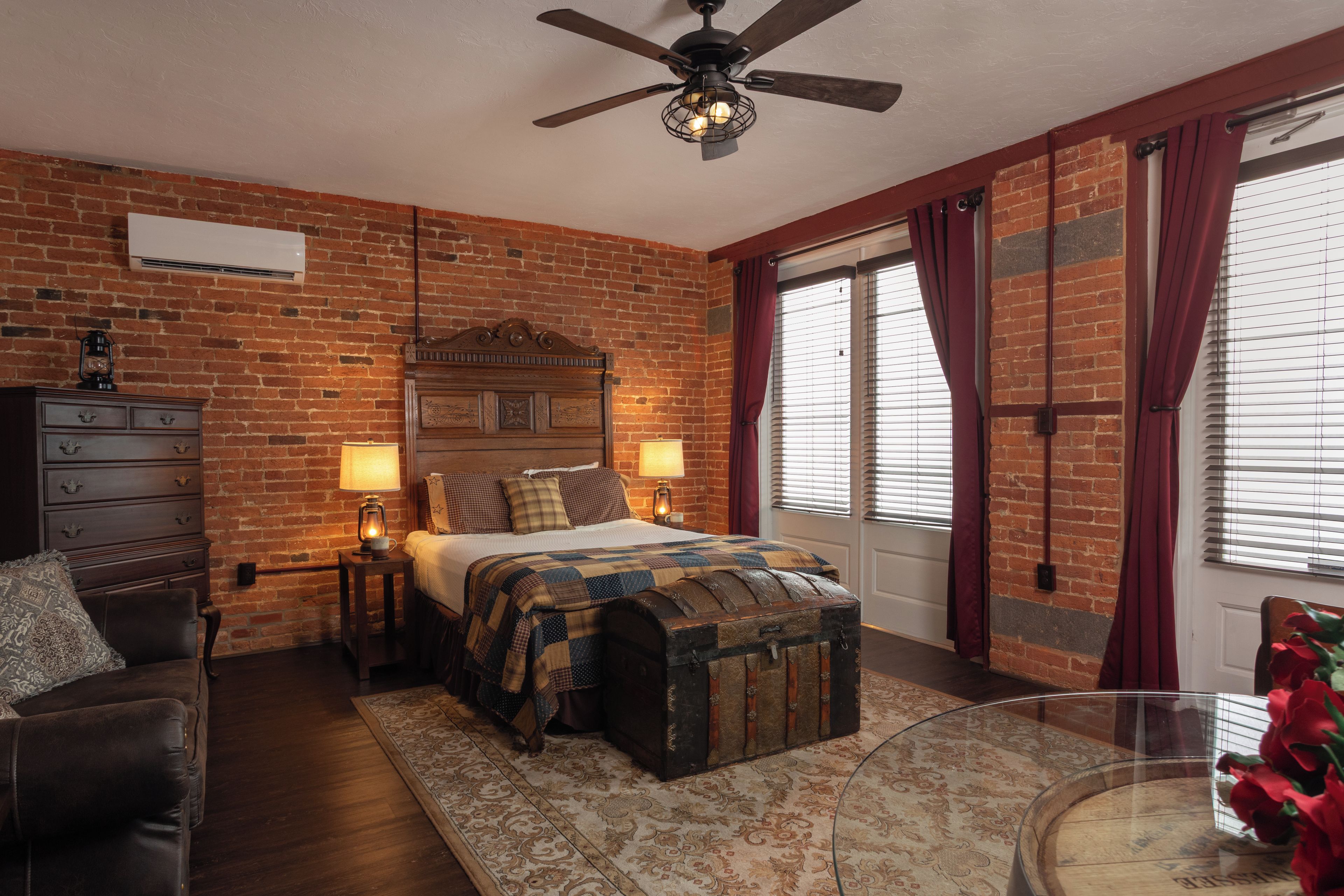 A bedroom with a brick wall, a wooden headboard, a bed with a quilt, a nightstand with a lamp, a dresser, a trunk, and a window with curtains.