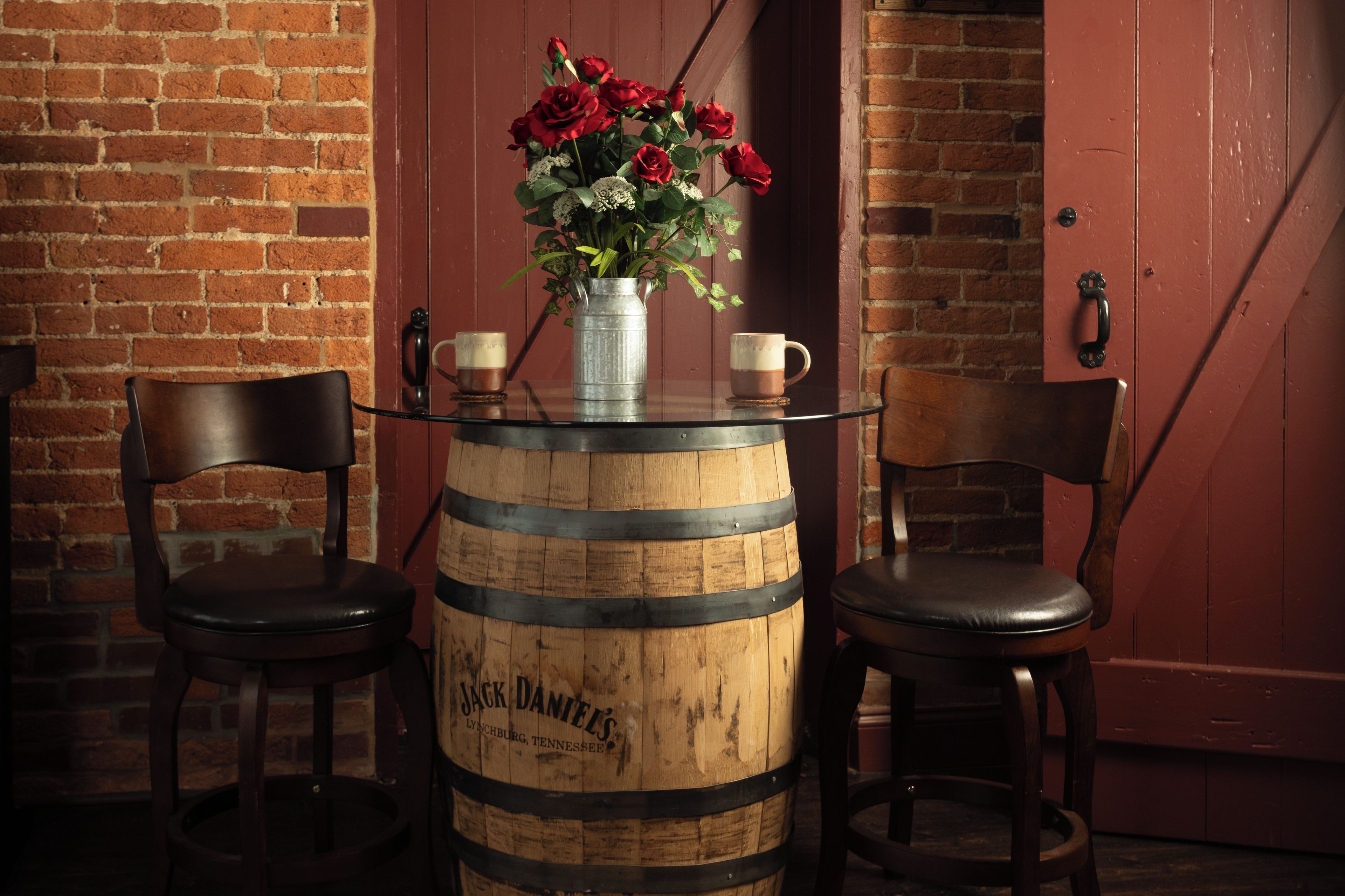 A rustic setting with a wooden barrel table, two chairs, a vase with red flowers, and a door in the background.
