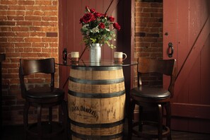 A rustic setting with a wooden barrel table, two chairs, a vase with red flowers, and a door in the background.