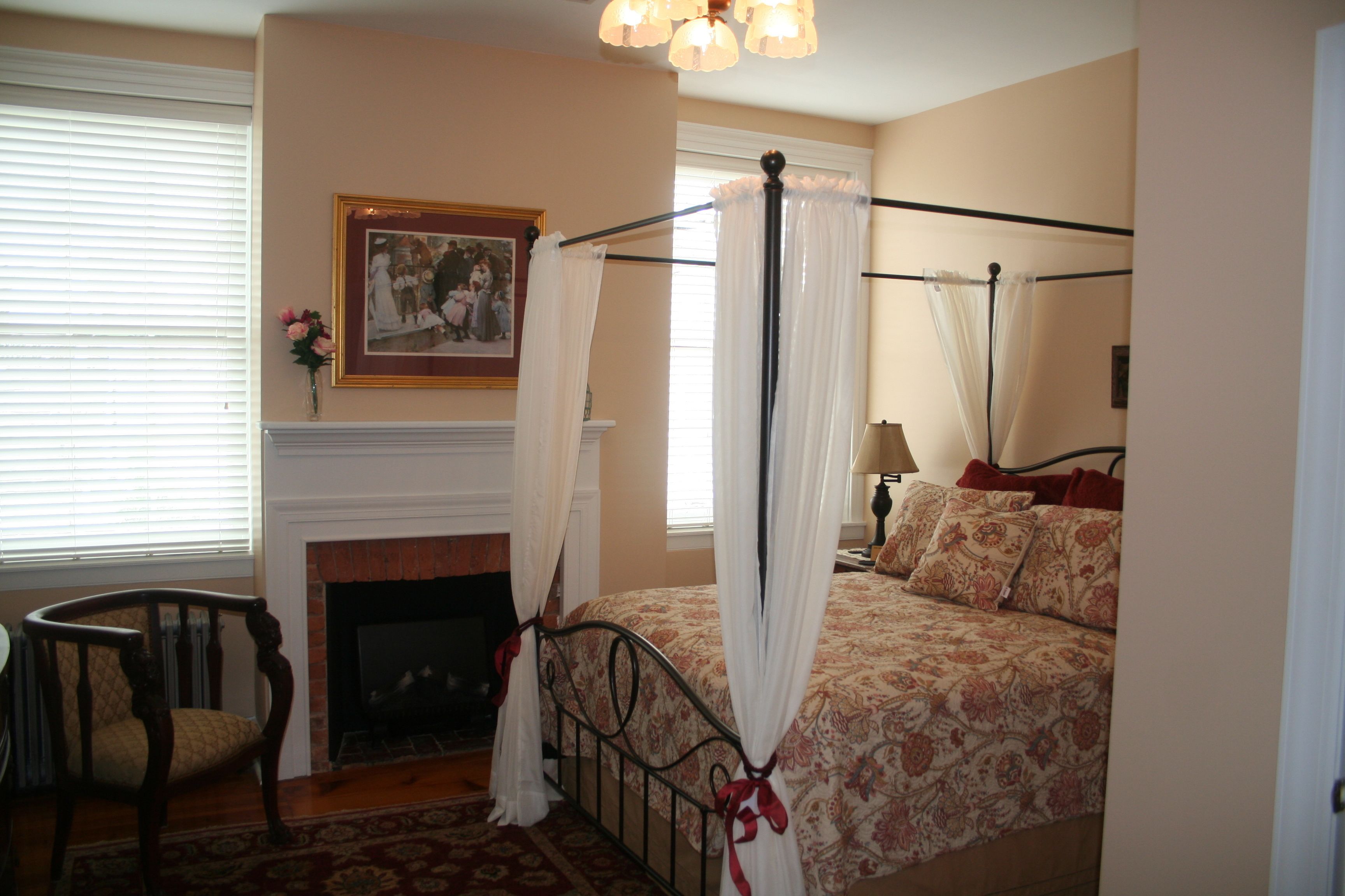 A bedroom with a four-poster bed, a fireplace, a chair, and a painting on the wall.