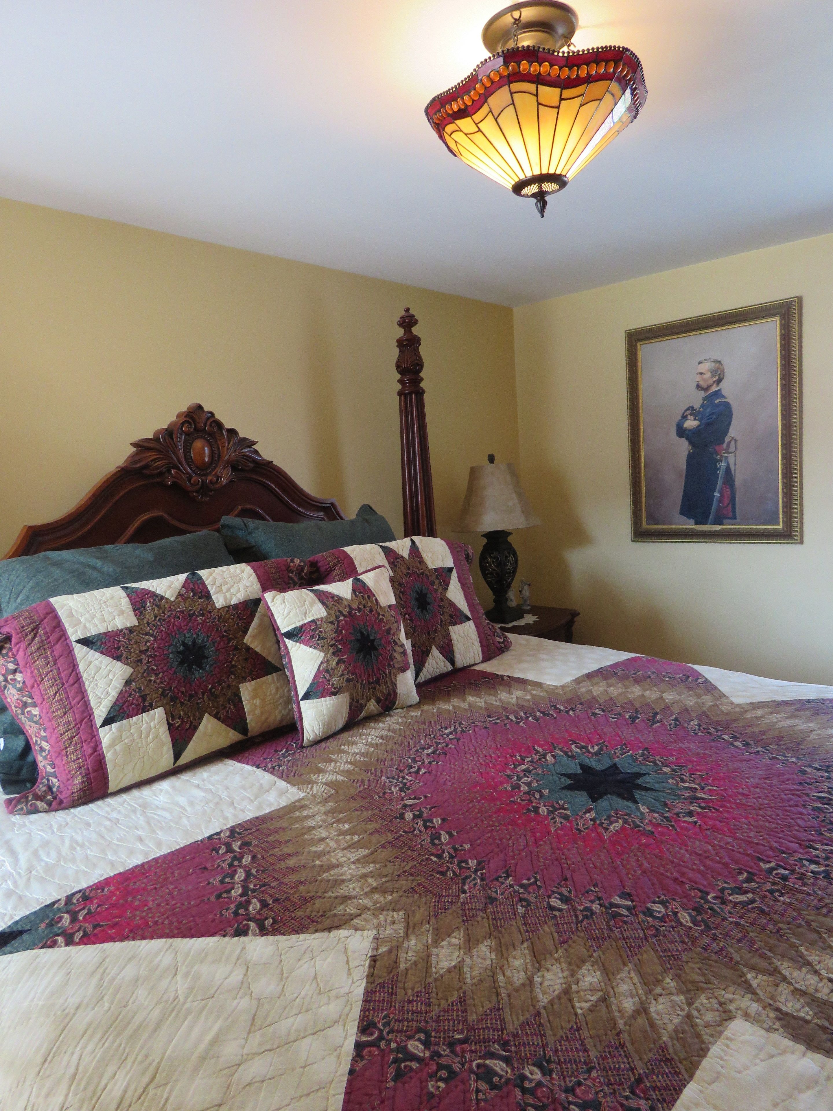 A bedroom with a large bed, a decorative ceiling light, a framed portrait on the wall, and a nightstand with a lamp.