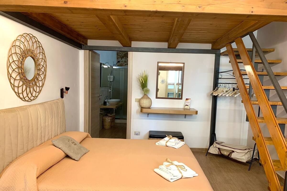 A bedroom with a bed, a wooden ladder, a bathroom visible through an open door, and a mirror on the wall.