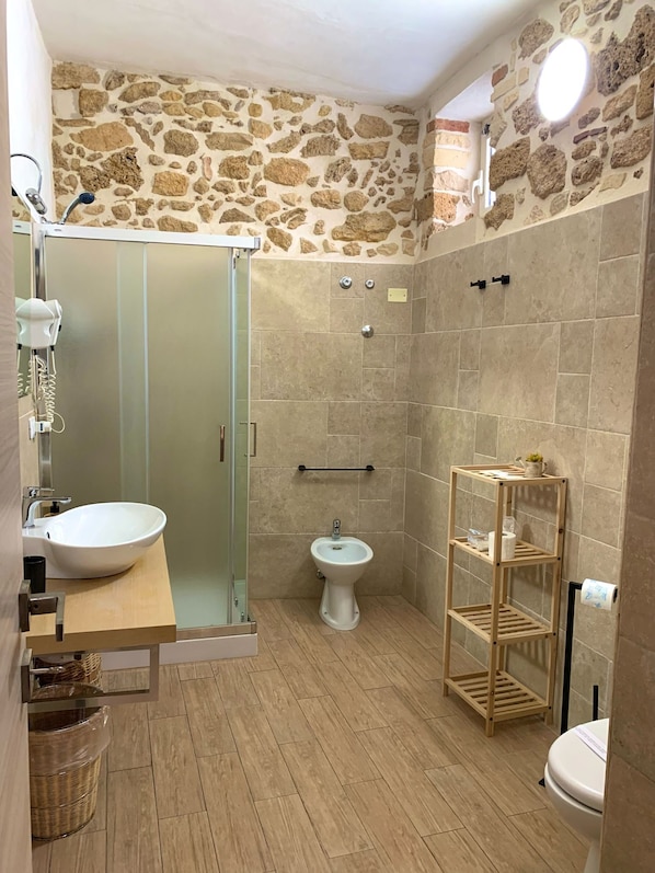 A bathroom with a stone wall, a glass shower enclosure, a sink, a bidet, and a wooden shelf.