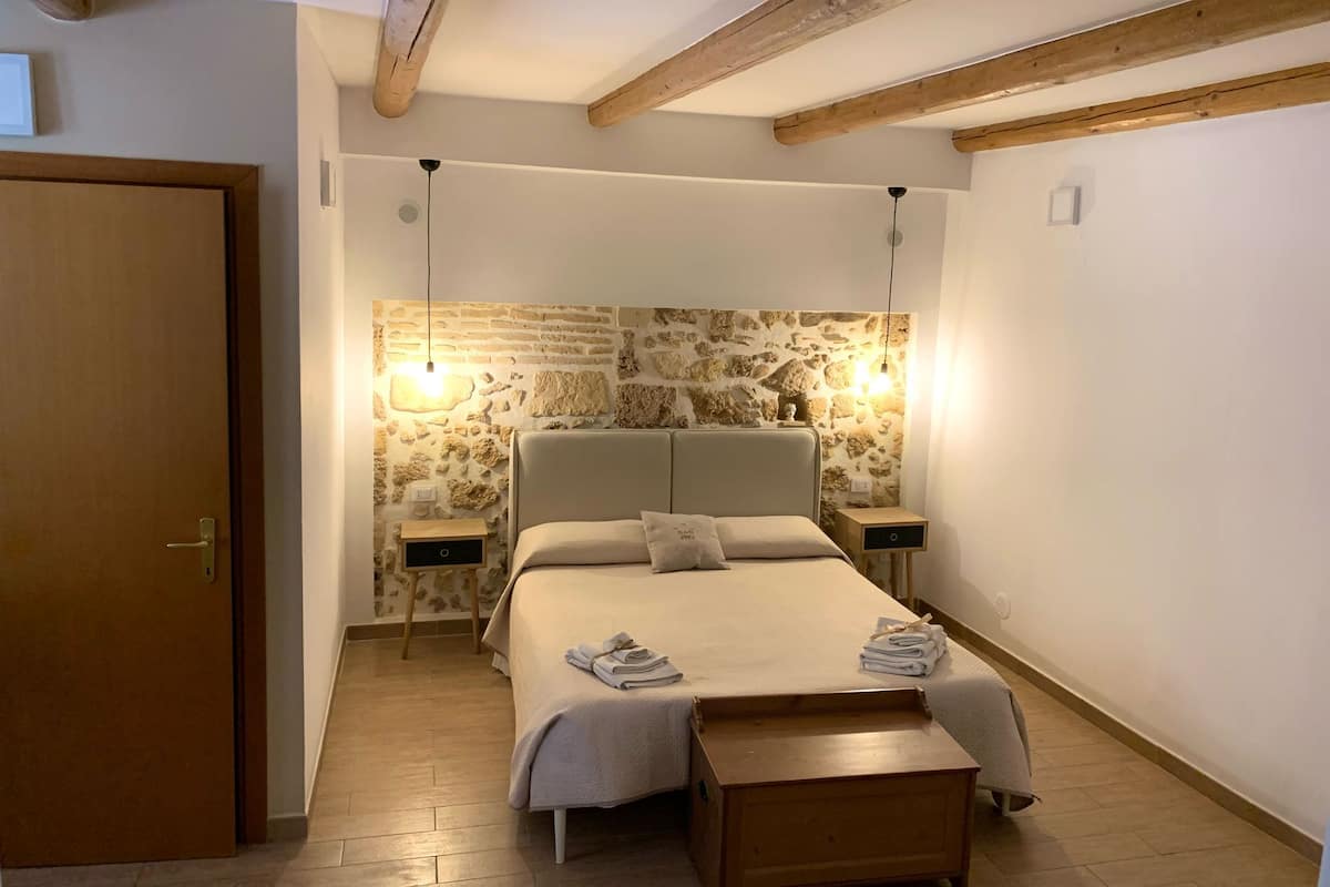 A bedroom with a stone wall, a bed with a wooden headboard, two bedside tables, and a wooden bench.