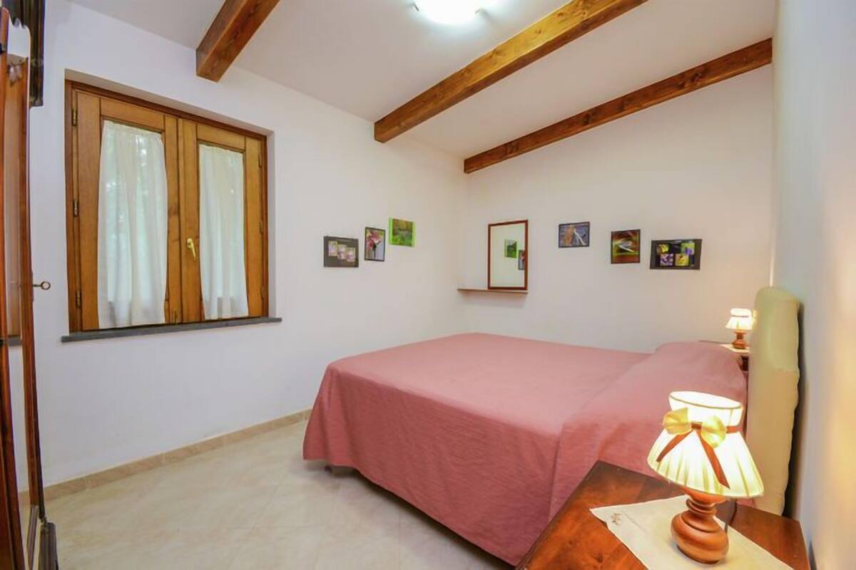 A bedroom with a single bed, wooden beams on the ceiling, a window with curtains, and a bedside lamp.