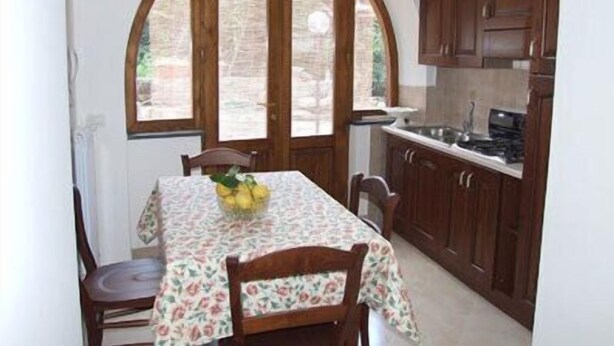 Een keuken met houten kastjes, een eettafel met een tafelkleed met bloemenprint en een raam met uitzicht op groen.