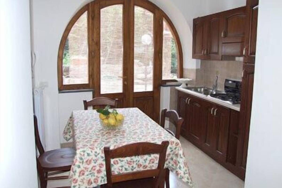 A kitchen with wooden cabinets, a dining table with a floral tablecloth, and a window with a view of greenery.