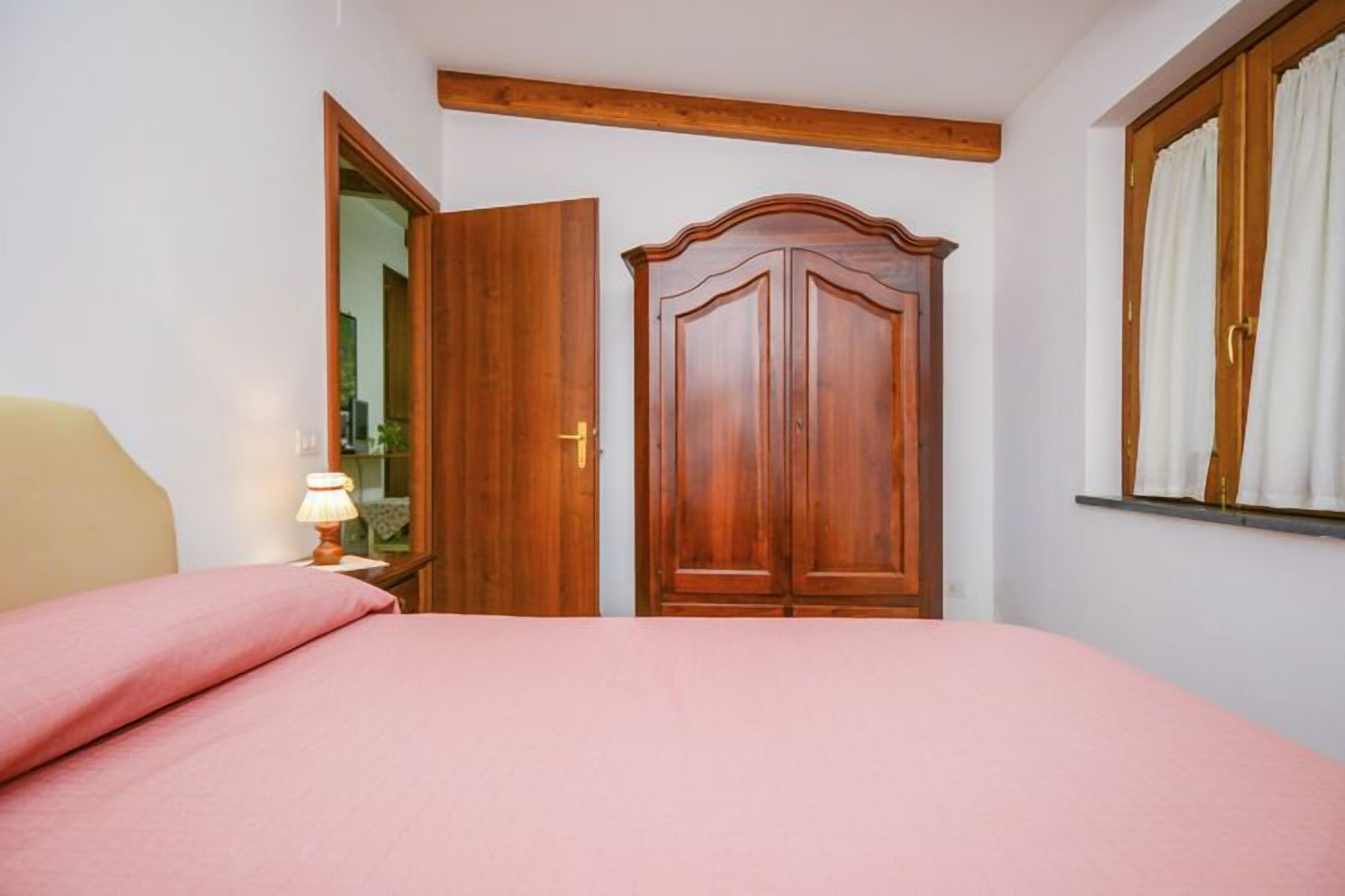 A bedroom with a bed, a wooden wardrobe, a nightstand with a lamp, and a window with curtains.