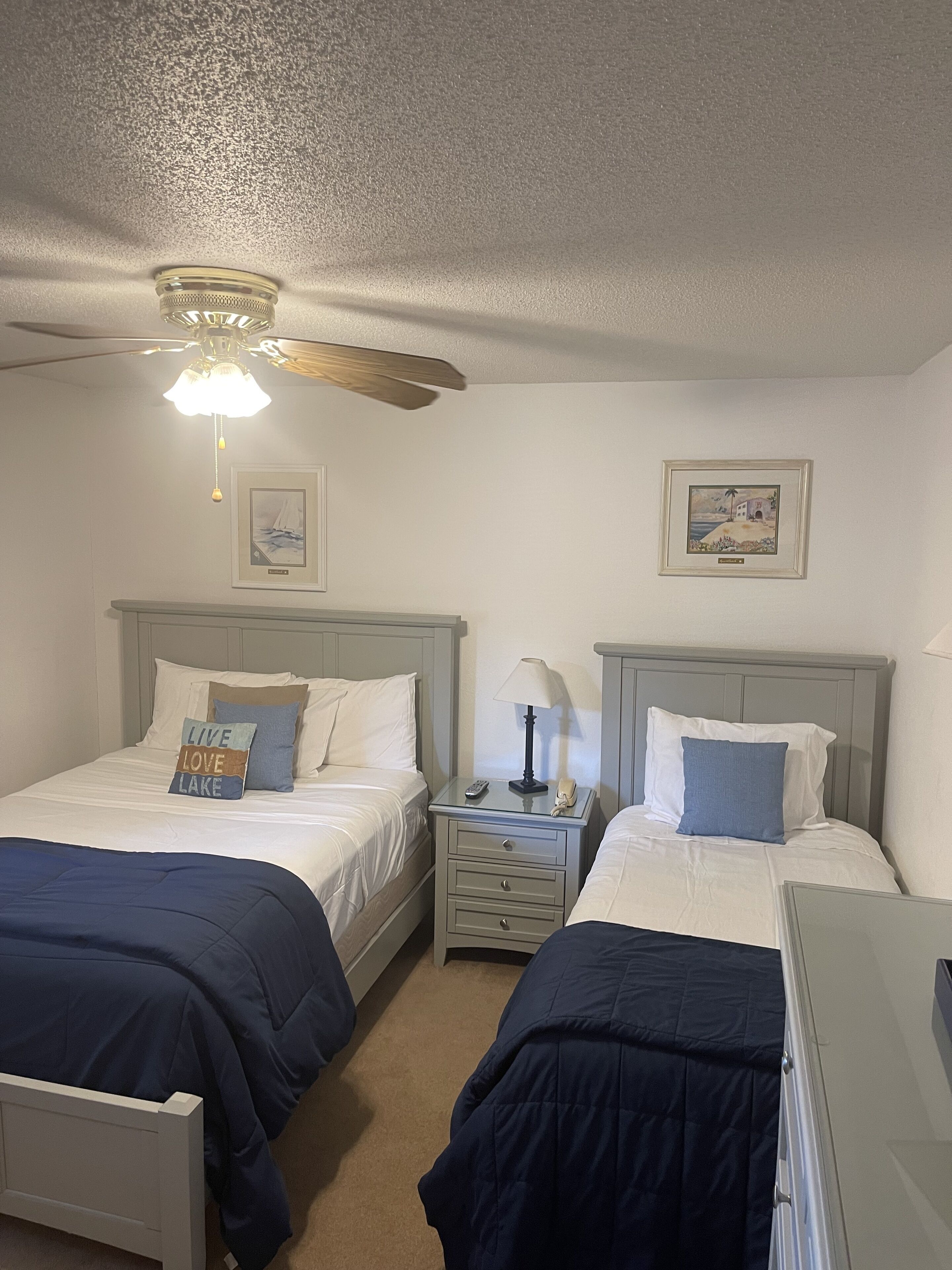 A hotel room with two beds, a ceiling fan, a nightstand with a lamp, and framed pictures on the wall.
