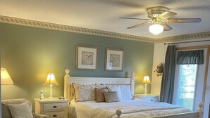 Une chambre à coucher comprenant un lit, une chaise, deux tables de chevet, un ventilateur de plafond et des œuvres d’art encadrées accrochées au mur.