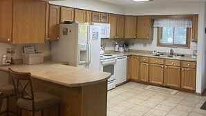 Une cuisine avec des meubles en bois, un îlot central doté de tabourets, un réfrigérateur, une cuisinière et une fenêtre avec des stores.