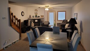 Un coin repas avec une table, des chaises et un ventilateur de plafond. On y trouve une cheminée agrémentée d’objets décoratifs, et un téléviseur est visible au fond.