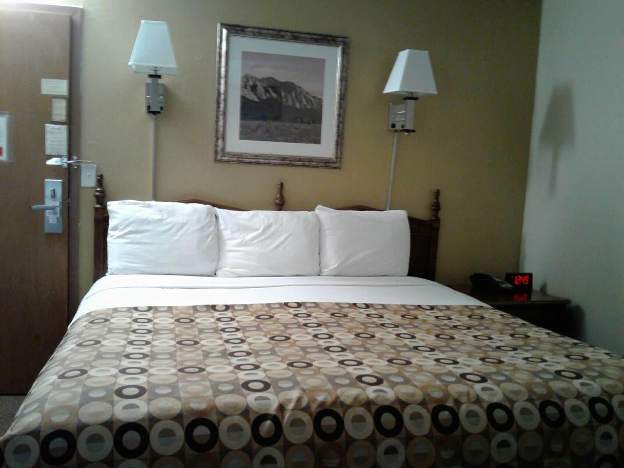 Una camera d'albergo con un letto, due lampade sui comodini, un quadro appeso alla parete, un comodino con una sveglia e una porta con serratura.