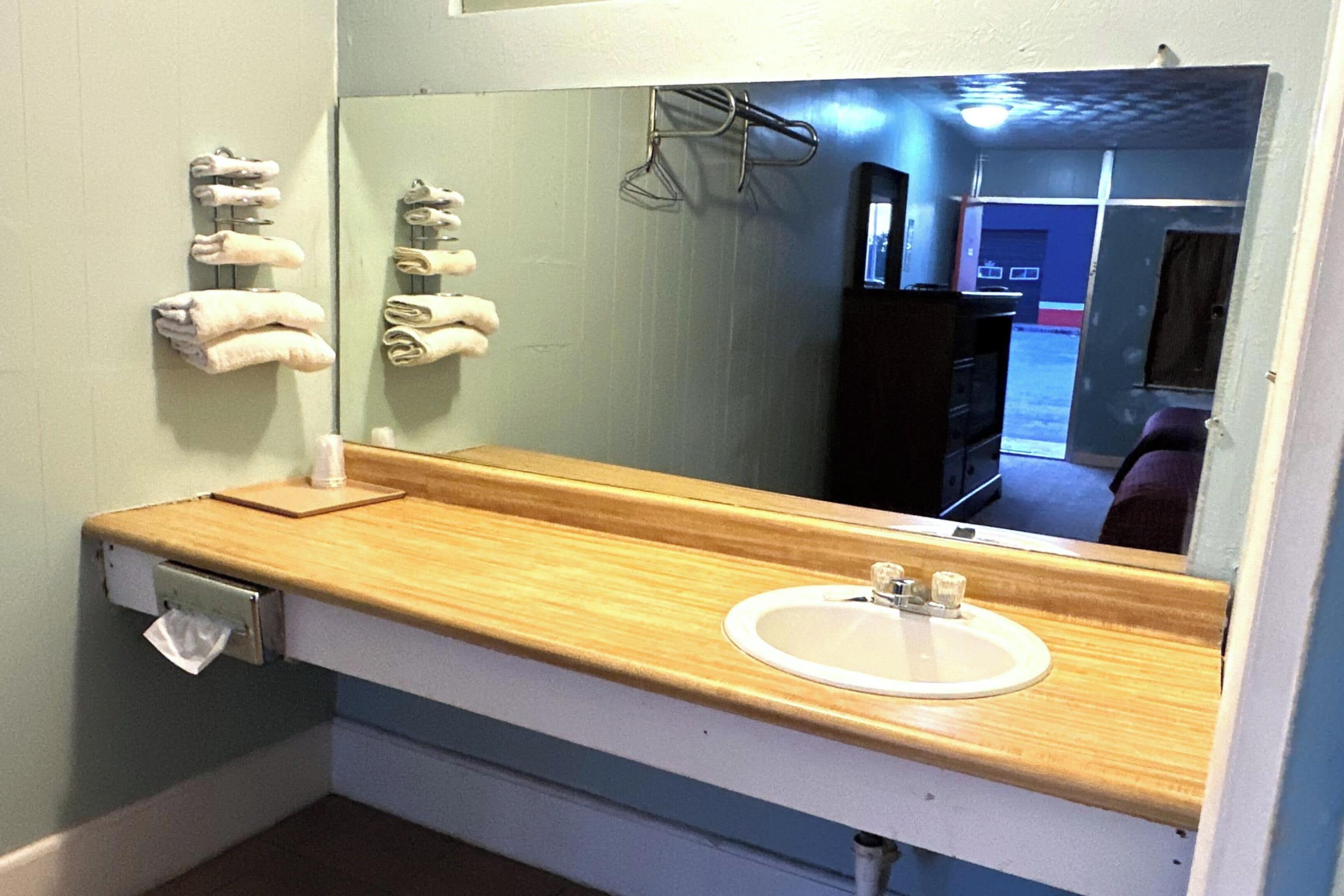 A bathroom with a wooden vanity, a white sink, and a mirror reflecting a door and a hallway.