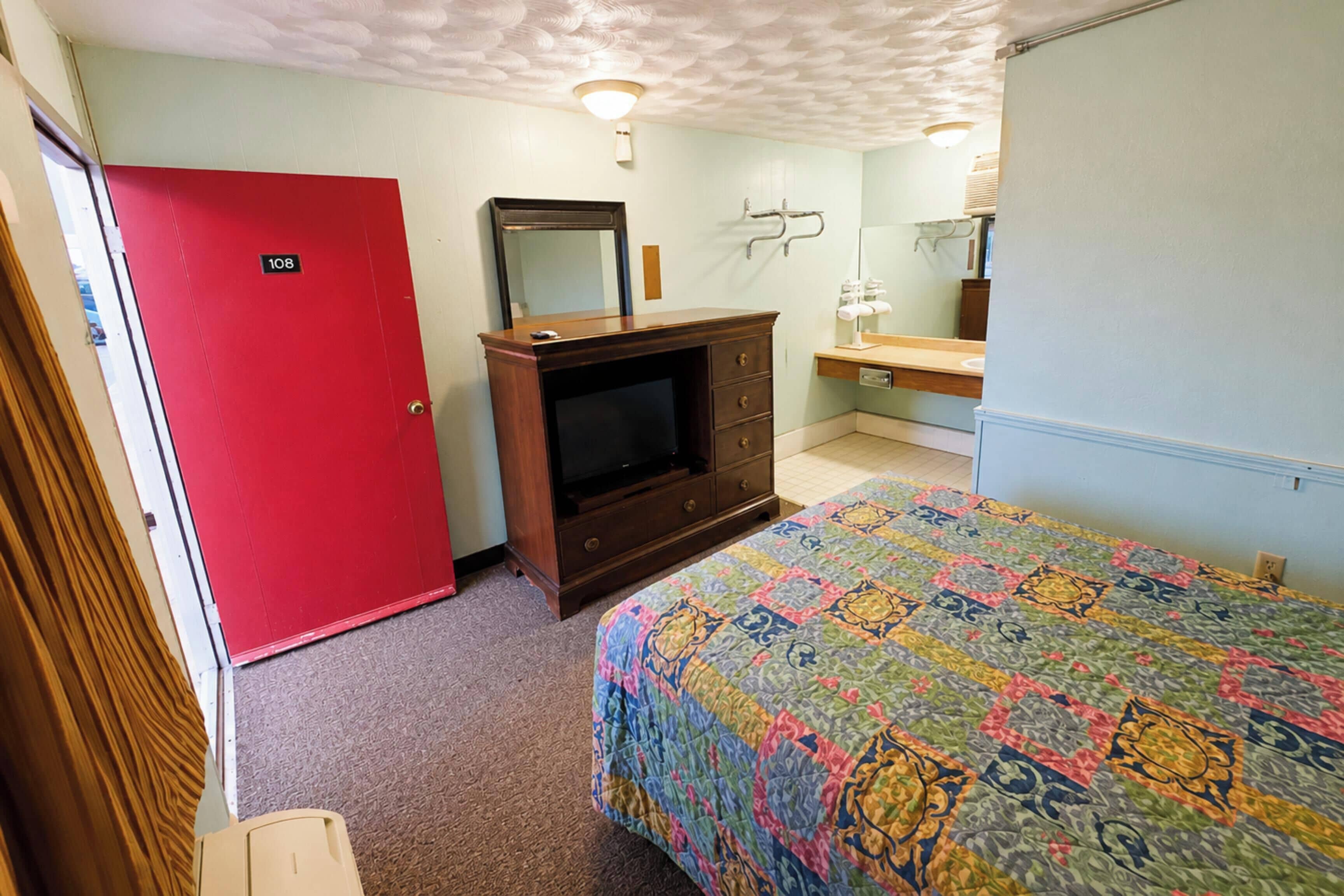 A room with a red door, a wooden dresser with a television, a bed with a colorful quilt, and a bathroom with a mirror and sink.