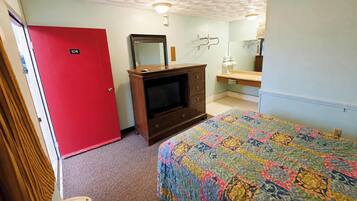 A room with a red door, a wooden dresser with a television, a bed with a colorful quilt, and a bathroom with a mirror and sink.