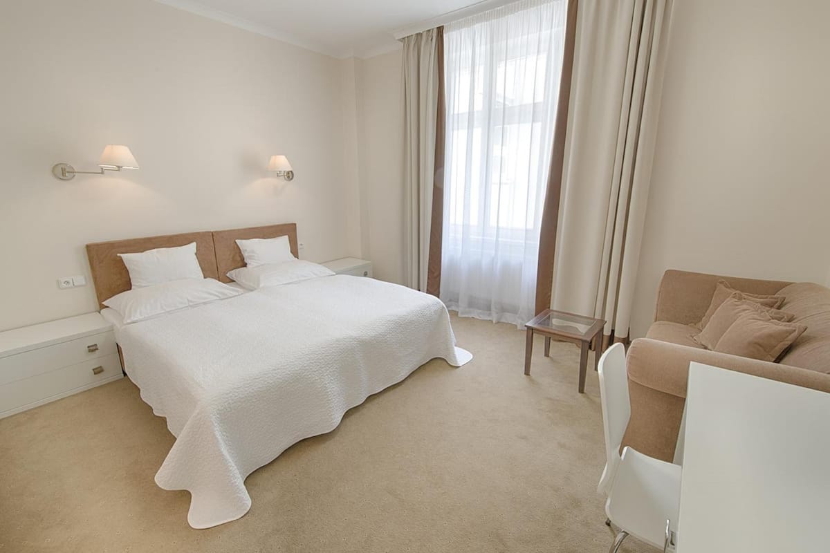 Habitación de hotel con una cama grande, una mesita pequeña, un sofá y una ventana con cortinas.