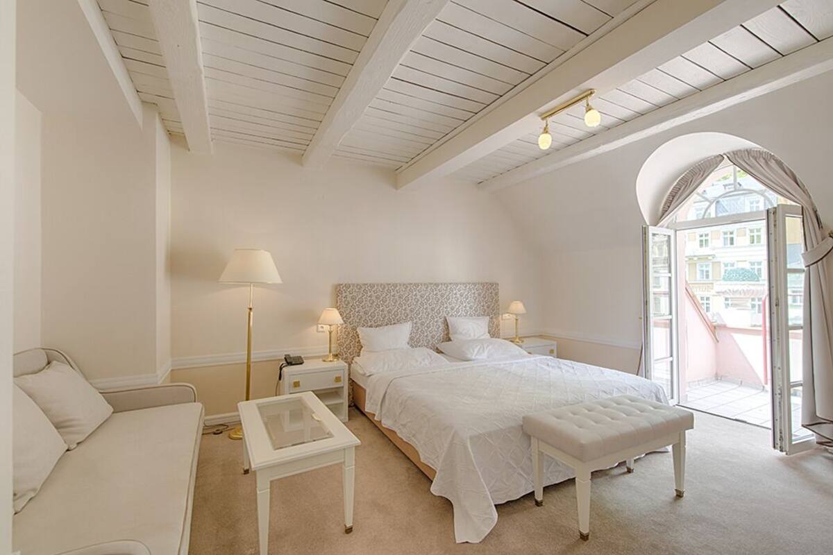 Un dormitorio con cama, mesita de noche, banco y ventana con cortinas.