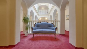 A hallway with a red carpet, a blue sofa, and white arches. - Hotel Embassy (Karlovy Vary)