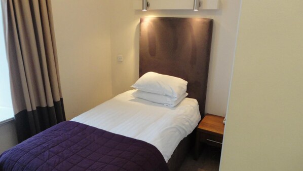 Un lit bien fait, avec du linge de lit blanc et une couverture violette, une table de chevet en bois et un luminaire fixé au mur.