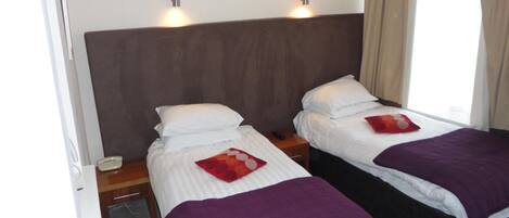 Ein Hotelzimmer mit zwei Einzelbetten, jedes mit einer violetten Decke und einem roten Kissen. An den Nachttischen stehen Lampen und ein Telefon. Das Zimmer ist in neutralen Farben gehalten, mit einem braunen Kopfteil und weißen Wänden.