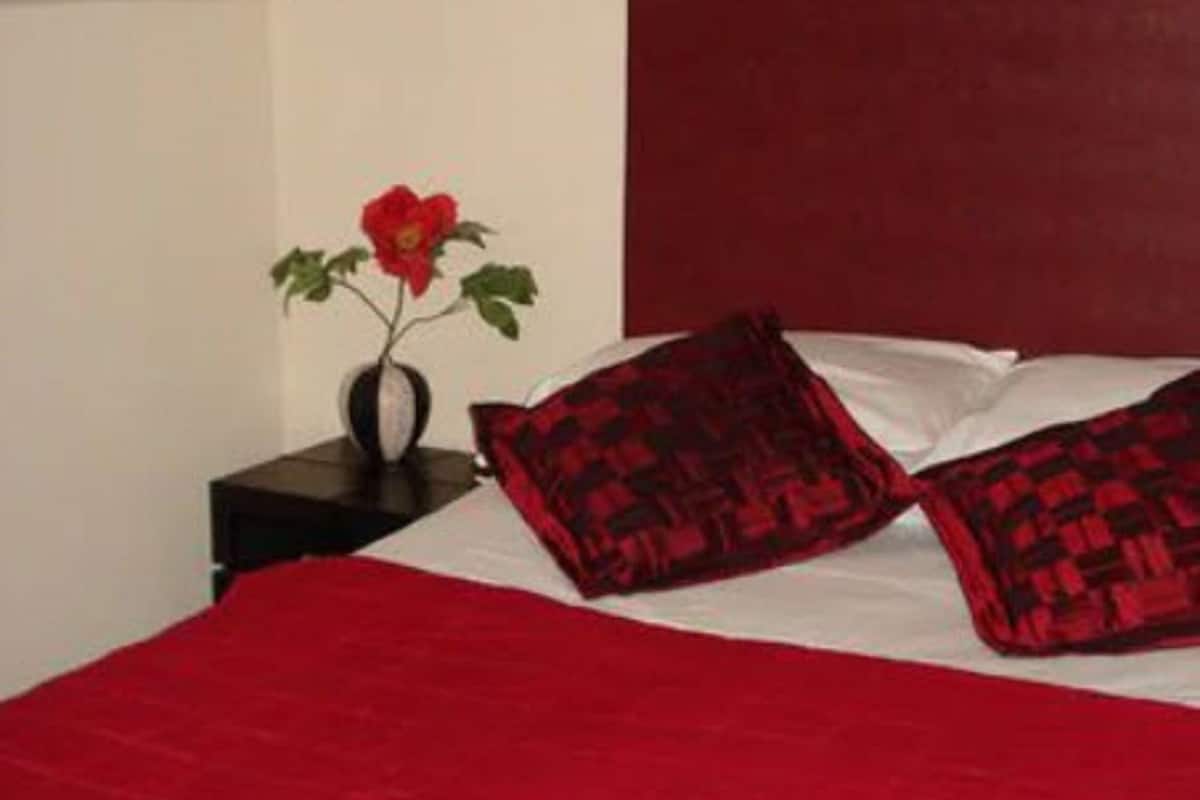 A bedroom with a red bedspread, two patterned pillows, a vase with a red flower, and a dark headboard.