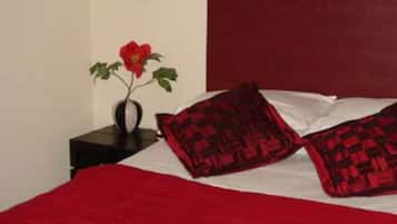 Ein Schlafzimmer mit roter Bettdecke, zwei gemusterten Kissen, einer Vase mit einer roten Blume und einem dunklen Kopfteil.