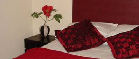 A bedroom with a red bedspread, two patterned pillows, a vase with a red flower, and a dark headboard.
