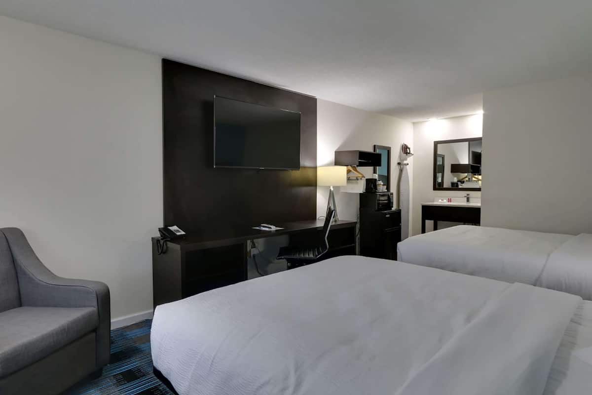 Habitación de hotel con dos camas, un escritorio, un televisor de pantalla plana, una silla, una lámpara y un espejo.
