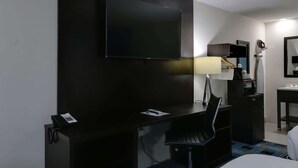 Hotelkamer met een donker houten bureau, een zwarte stoel, een flatscreen televisie aan de muur, een bureaulamp, een kleine koelkast, een spiegel en een bed.