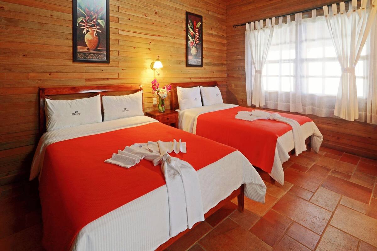 A room with two beds, wooden walls, a window with curtains, and a wall-mounted lamp.