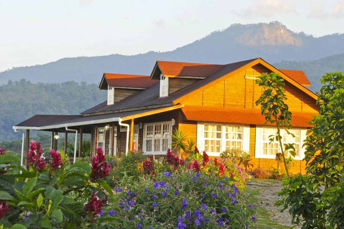 A wooden house with a covered porch, surrounded by colorful flowers and mountains in the background.