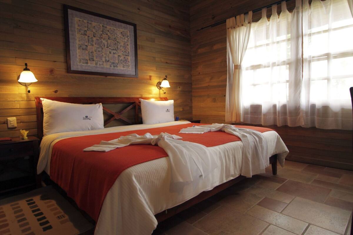 A wooden room with a bed, two bedside lamps, a framed picture, and a window with curtains.