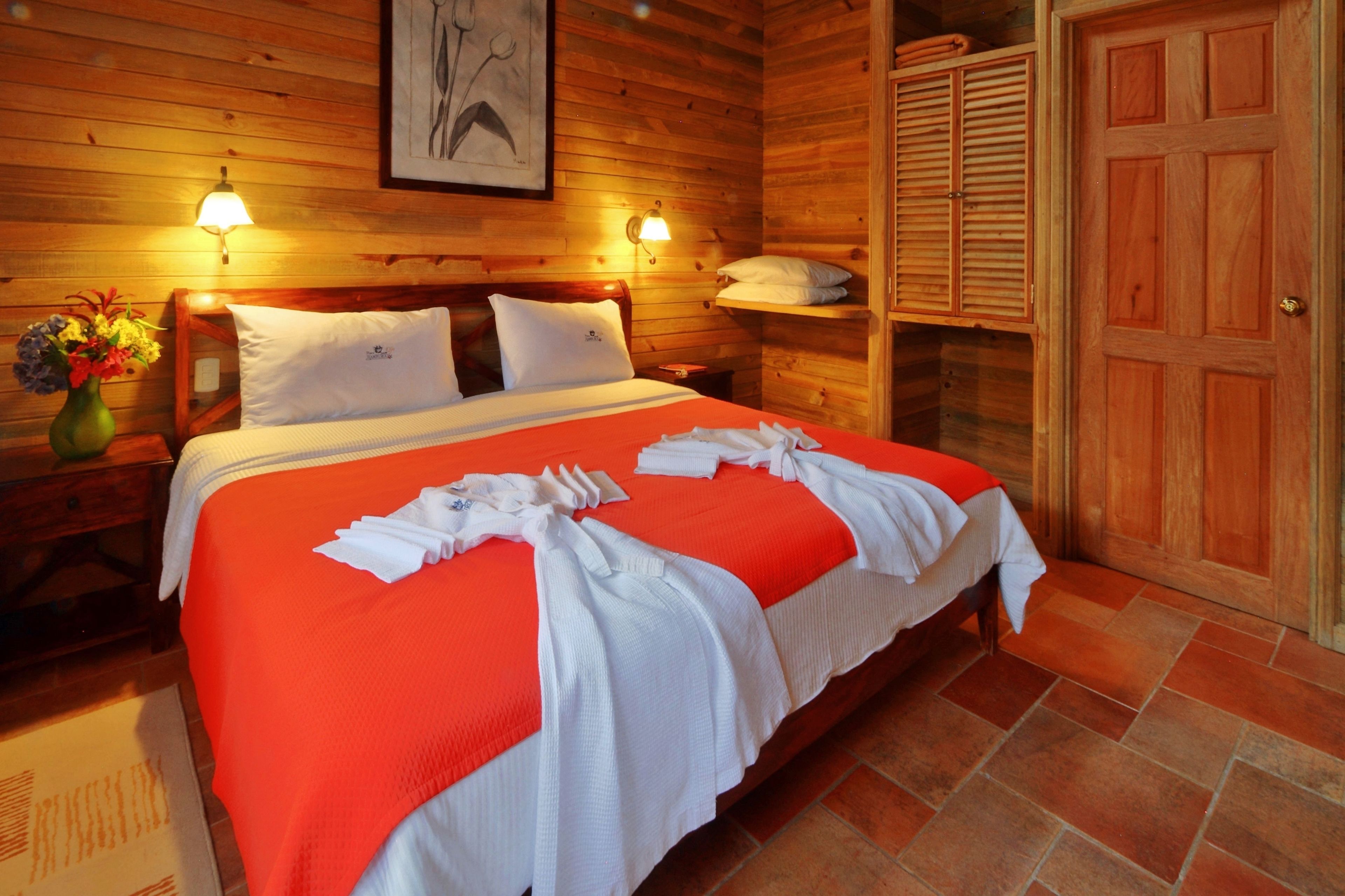 A wooden room with a bed, two wall-mounted lamps, a wooden wardrobe, and a framed picture on the wall.