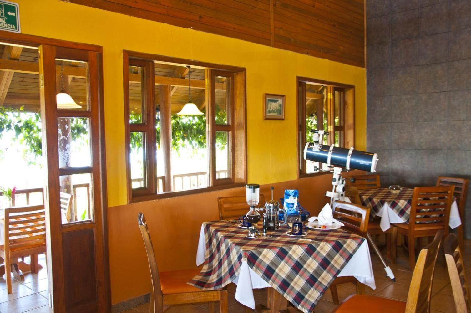 Comedor con mesas y sillas de madera, un telescopio y vistas al exterior con vegetación.