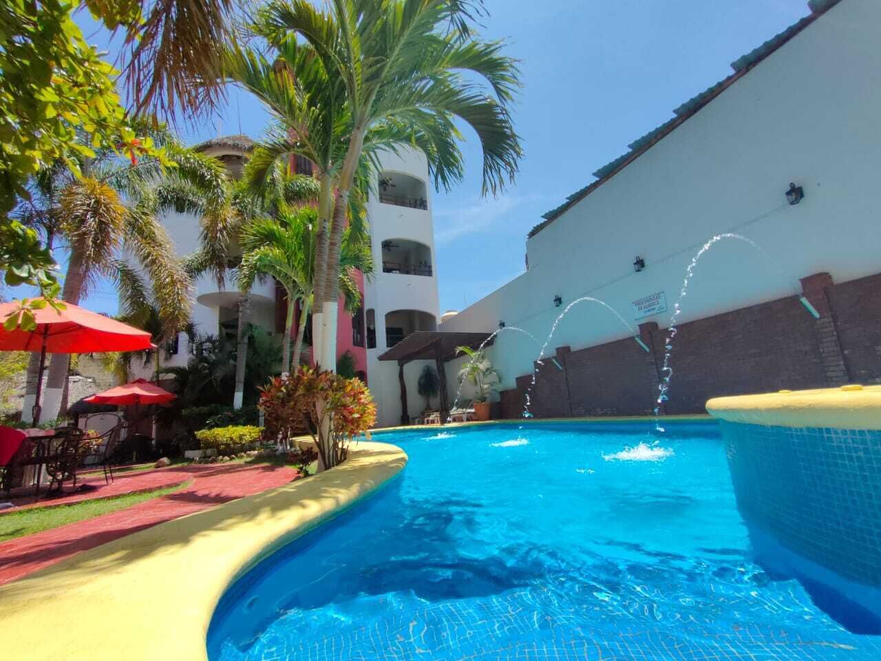 Piscina con una fuente de agua, rodeada de palmeras y sombrillas rojas.