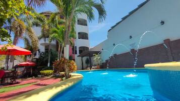 Piscina con una fuente de agua, rodeada de palmeras y sombrillas rojas.