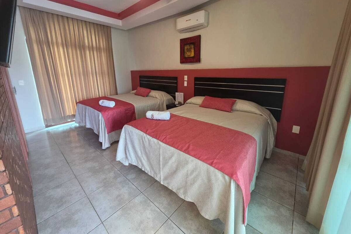 Habitación de hotel con dos camas, ventilador de techo, equipo de aire acondicionado y ventana con cortinas.