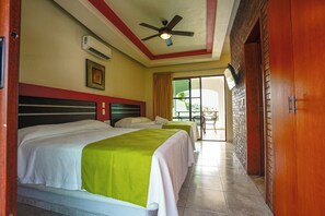 Quarto de hotel com duas camas, um ventilador de teto, aparelho de ar condicionado, parede de tijolos e uma porta de vidro deslizante para uma área externa.