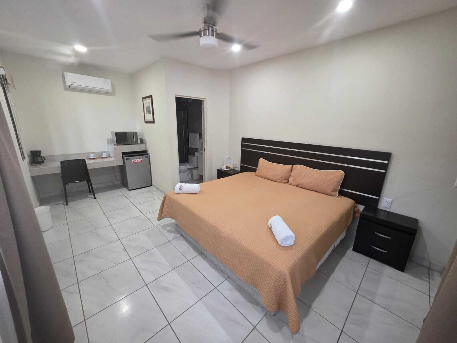 A hotel room with a bed, a desk, a chair, a small cabinet, and a ceiling fan.