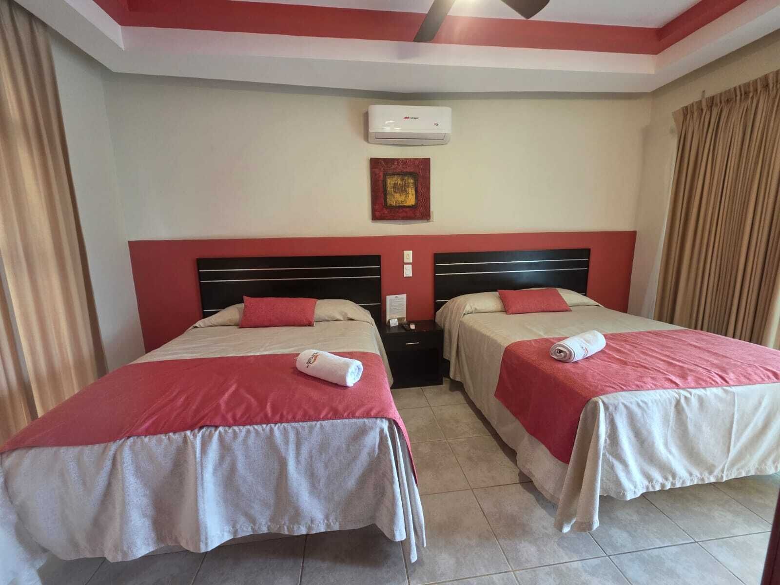 Habitación de hotel con dos camas, ventilador de techo, aire acondicionado y un cuadro en la pared.
