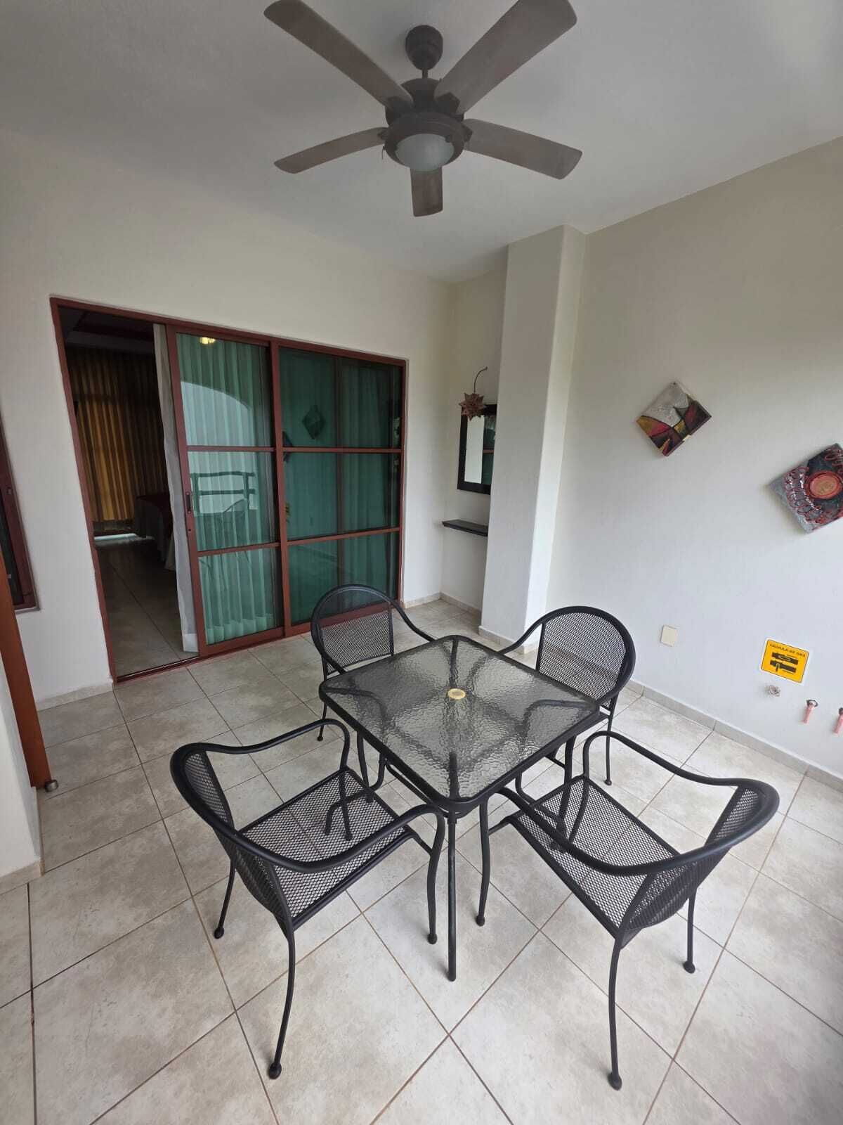 Una habitación con una mesa de vidrio, cuatro sillas, un ventilador de techo, un cristalero corredero y un pequeño estante con objetos.