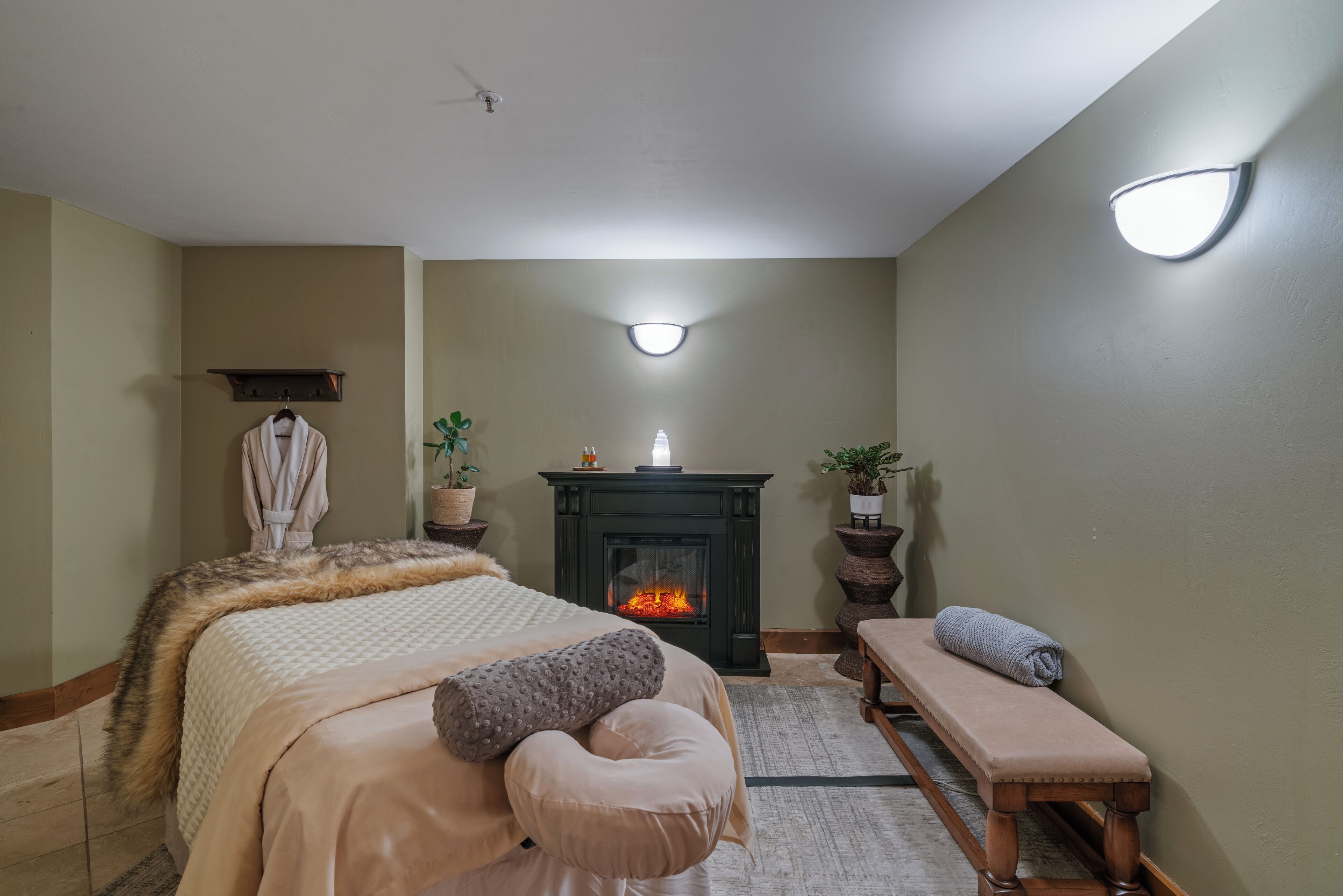 Un dormitorio con cama, banca, chimenea e iluminación en la pared.