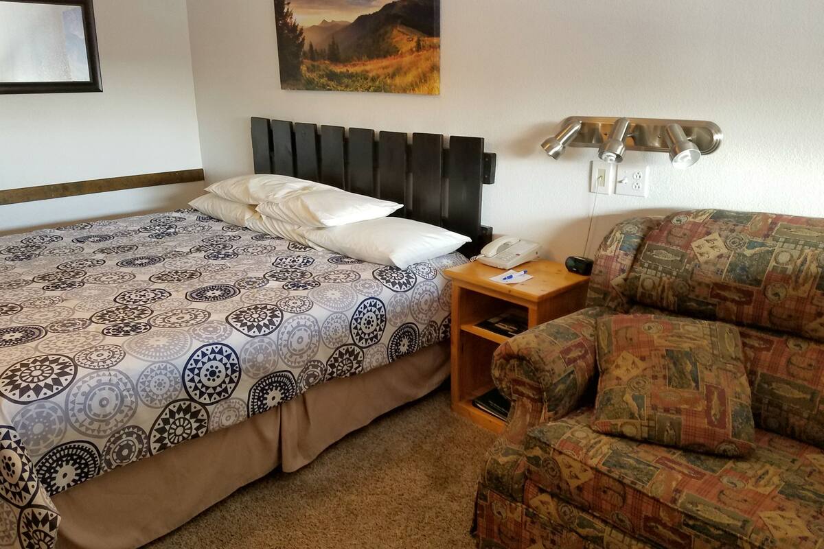 A hotel room with a bed, a nightstand with a phone, a lamp, a patterned sofa, and a painting on the wall.