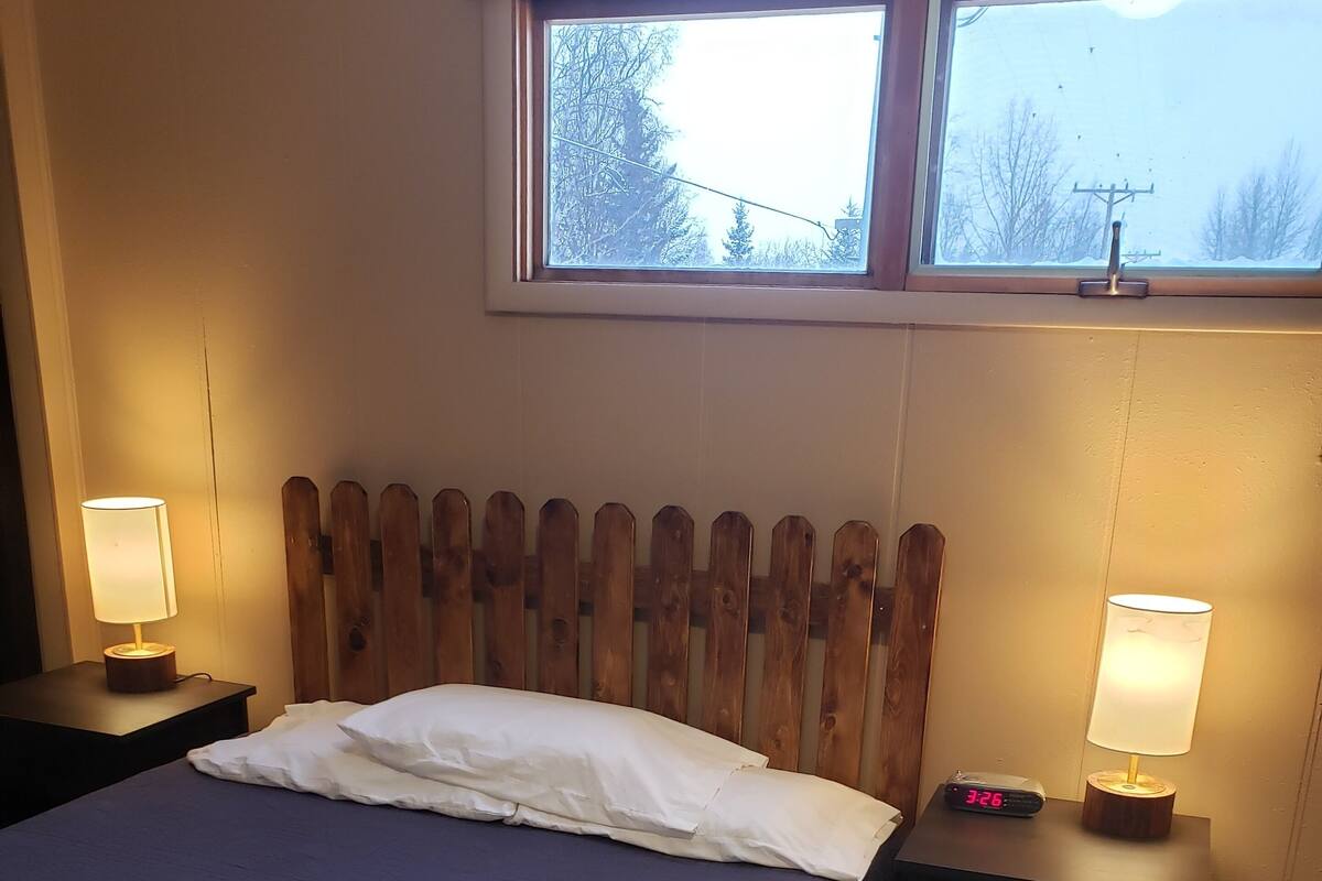A bedroom with a wooden headboard, a bed with white bedding, two bedside tables with lamps, and a window with a view of trees and a power line.