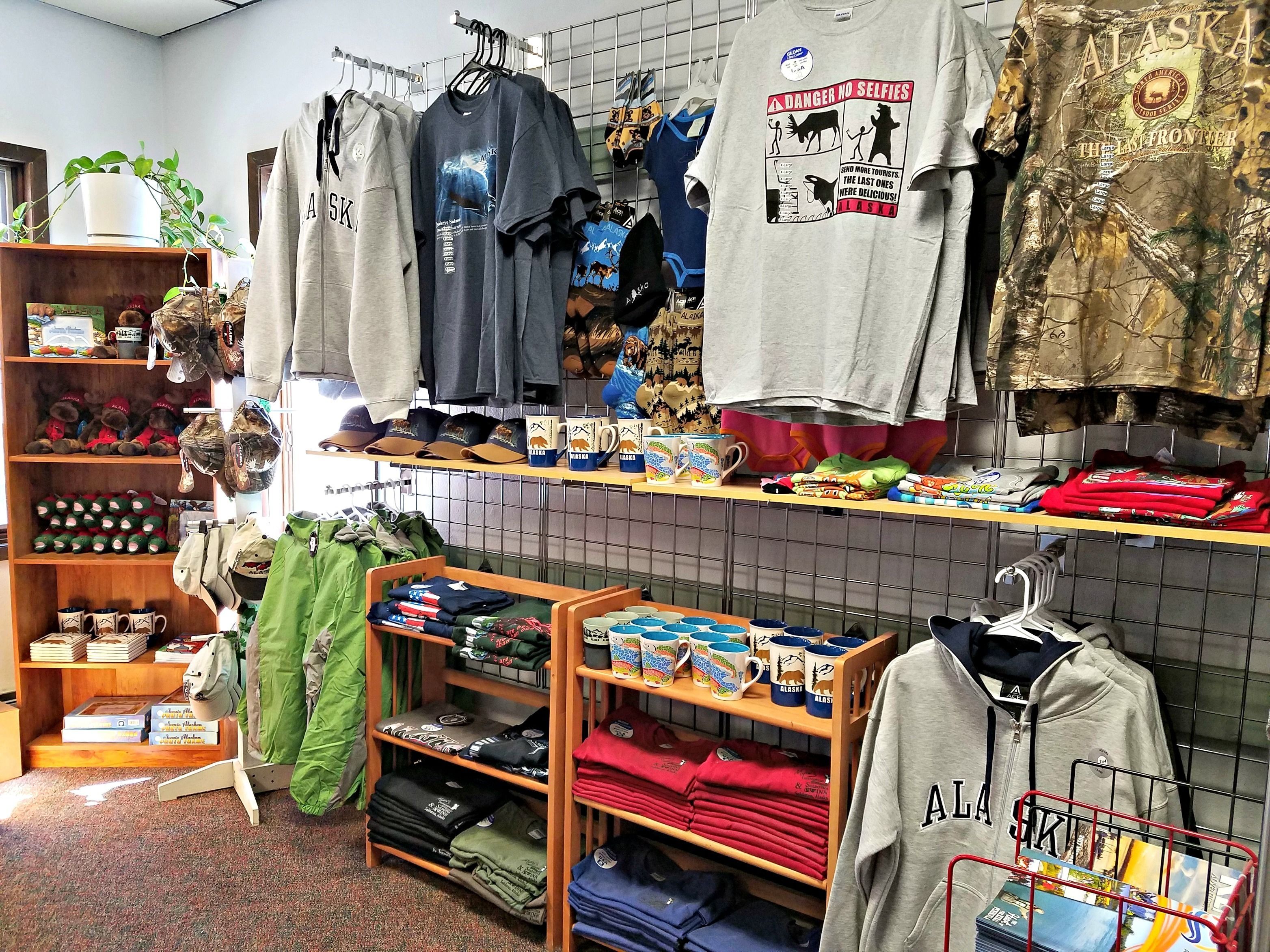 Una tienda de ropa con perchas llenas de camisetas, sudaderas y otras prendas, estantes con sombreros y tazas, y una exhibición de ropa doblada.